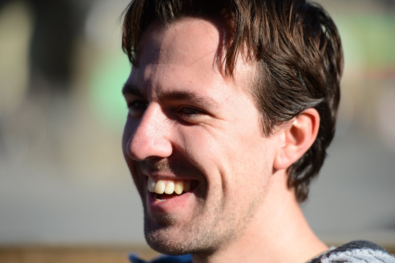 A man with short brown hair is smiling while looking to the side in bright sunlight.