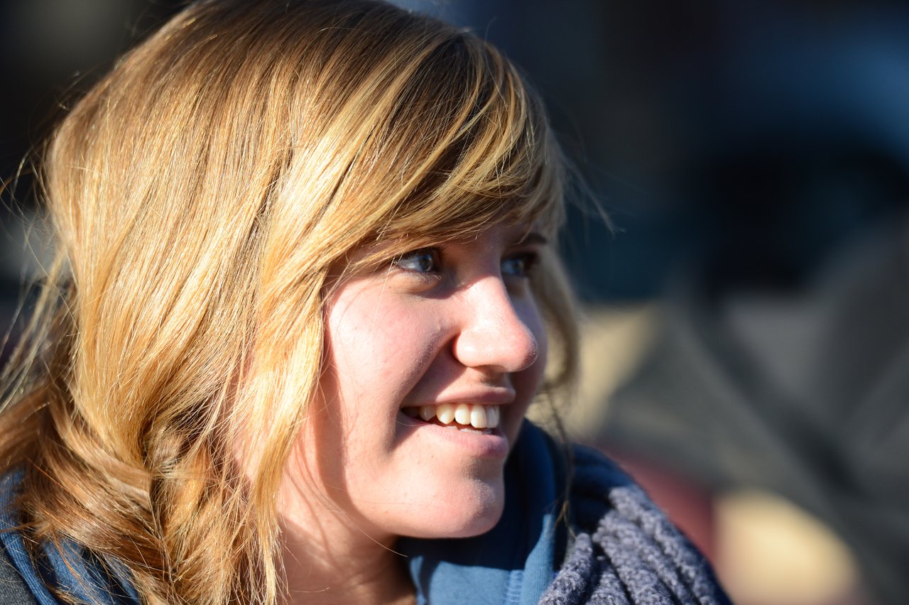 A person with blonde hair smiles while looking to the side in natural sunlight.