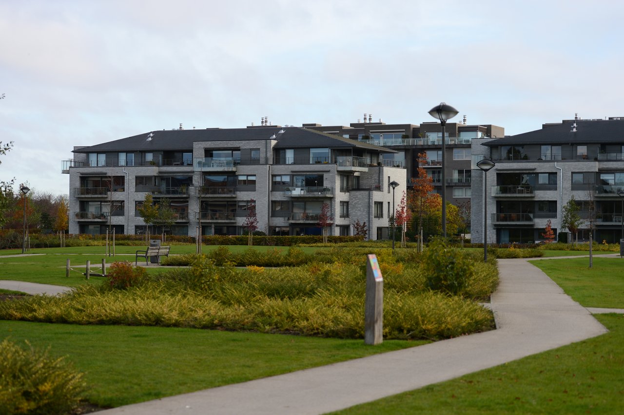 Modern apartment buildings with balconies surrounded by a landscaped park with pathways, benches, and street lamps.