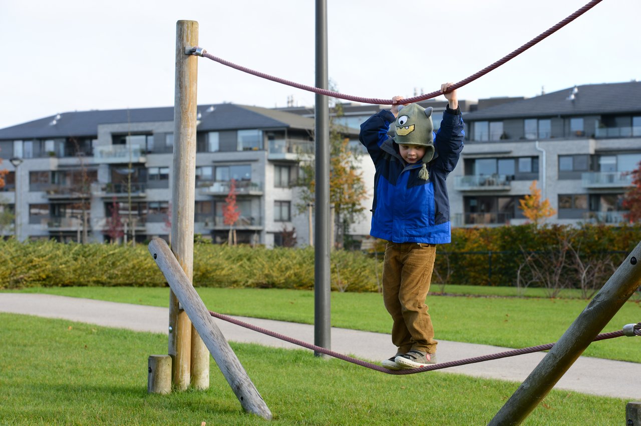 A child in a blue jacket and monster hat balances on a rope bridge at a playground.