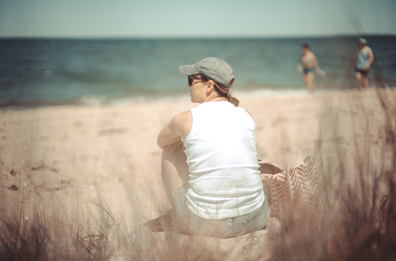 A woman in a white tank top and cap sits on the beach, looking toward the ocean.