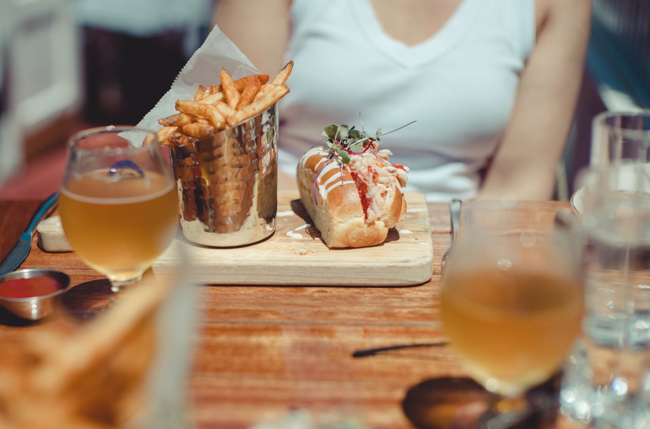 A lobster roll with fries and a drink on a wooden table at an outdoor restaurant.
