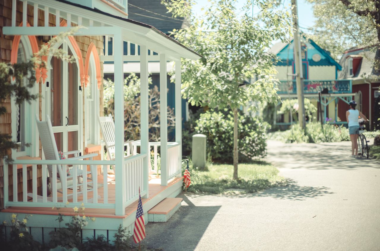 A colorful house with a porch and rocking chairs, while a person walks a bicycle down the street.