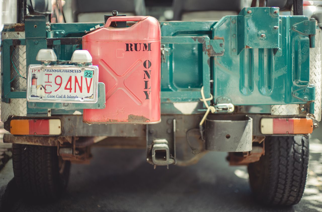 A red fuel can labeled "RUM ONLY" is attached to the back of a green Jeep with a Massachusetts plate.
