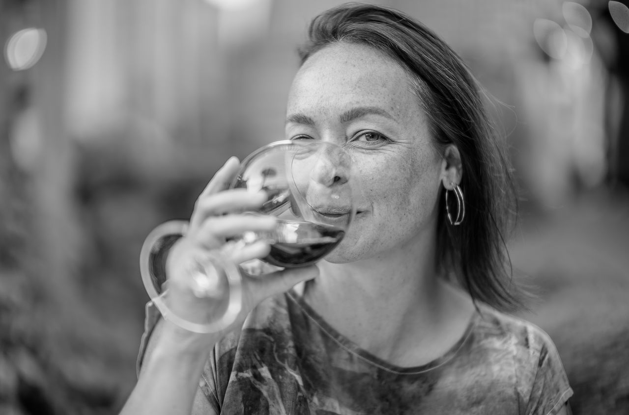 A woman winks while sipping red wine from a glass, holding it close to her face.