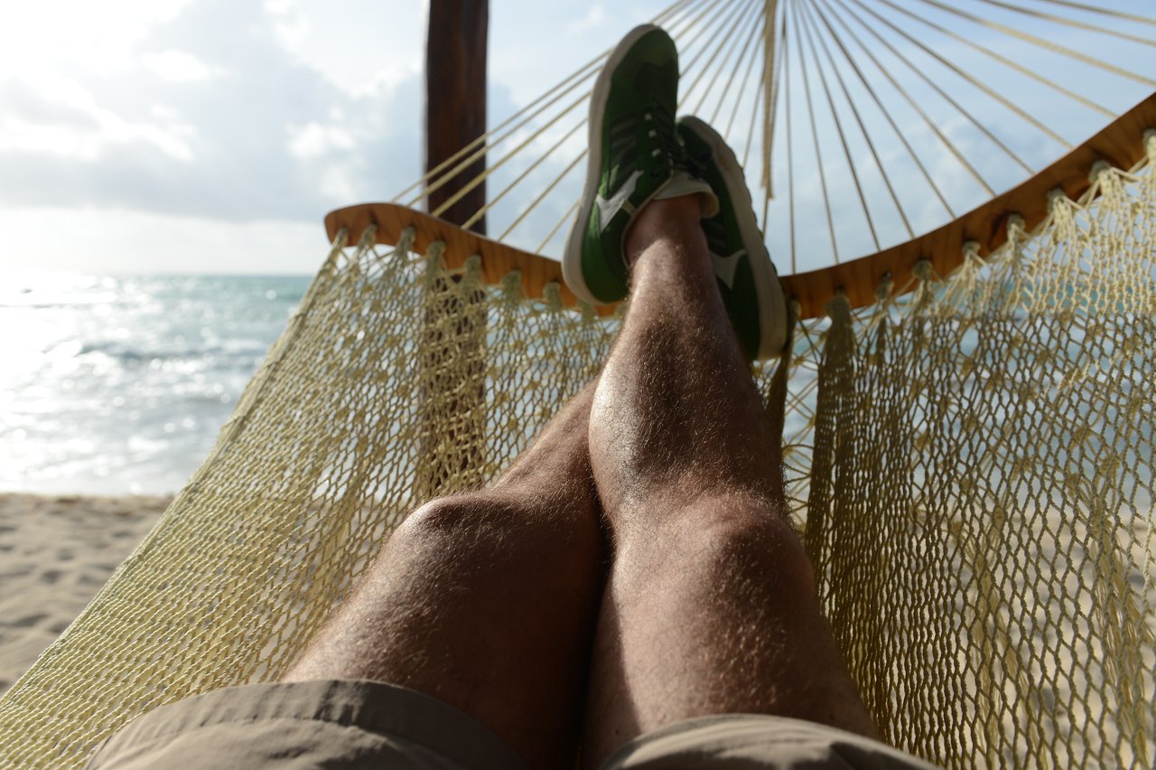 Person relaxing in a hammock with legs stretched out, facing the ocean on a sunny day.