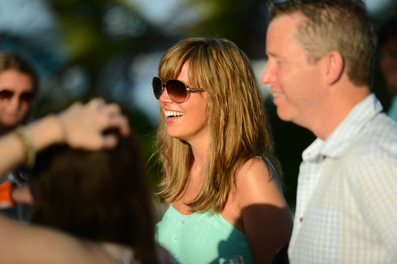 A woman in sunglasses and a light dress smiles while talking with others at an outdoor gathering.