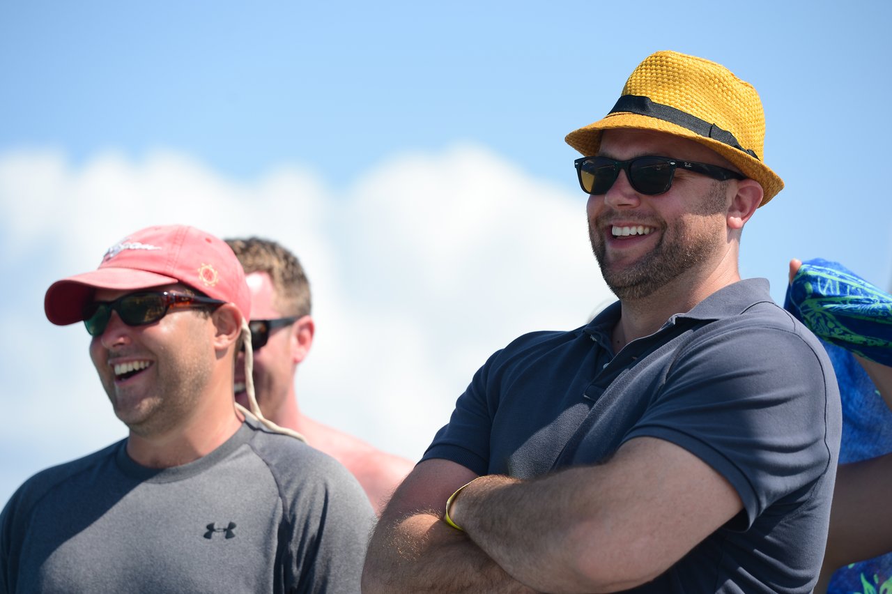 Two men wearing sunglasses and hats smile while looking at something off-camera under a bright sky.
