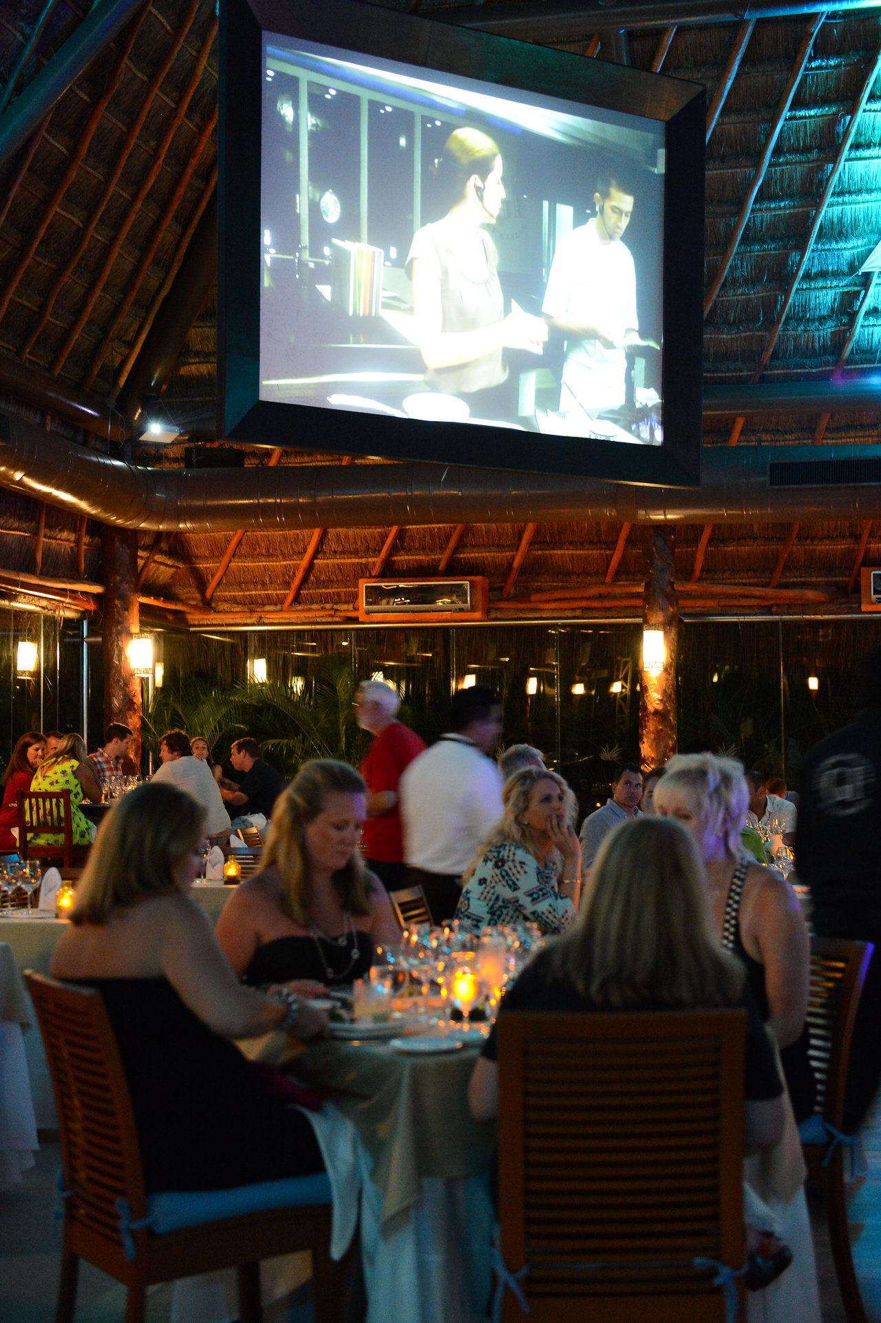 People dining at a restaurant with candlelit tables, while a large screen displays a cooking demonstration.