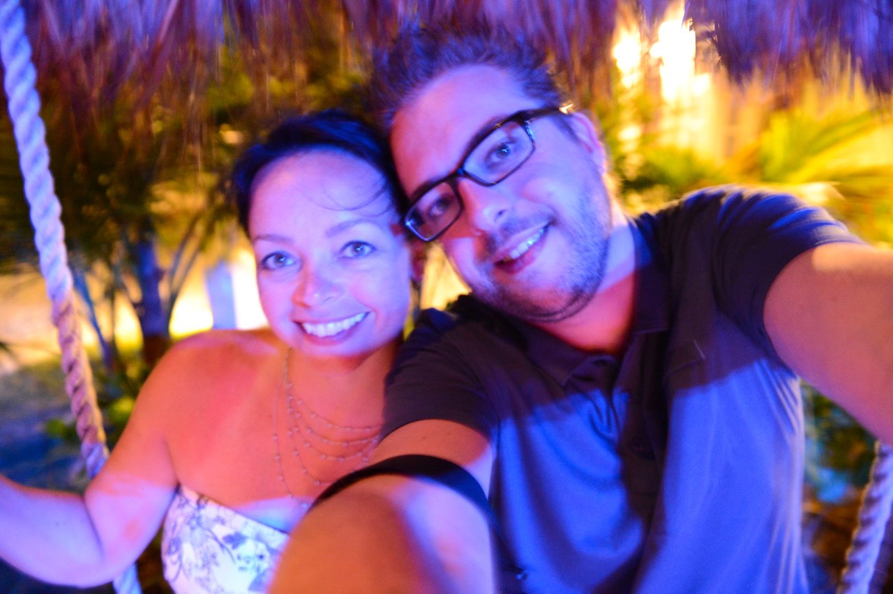 A smiling couple takes a selfie while sitting on a swing under a thatched roof at night.