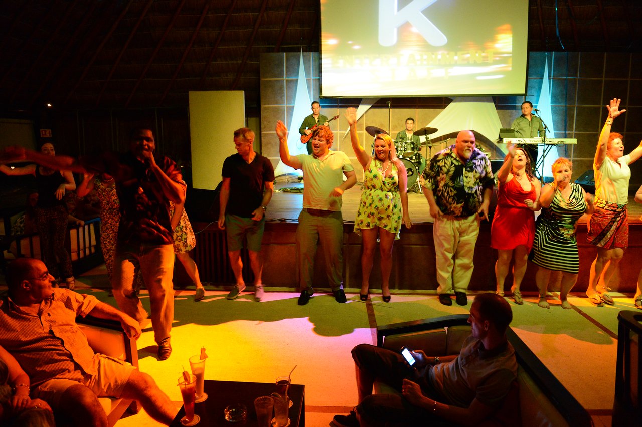 A group of people dances on stage while a live band performs in a lively indoor setting.