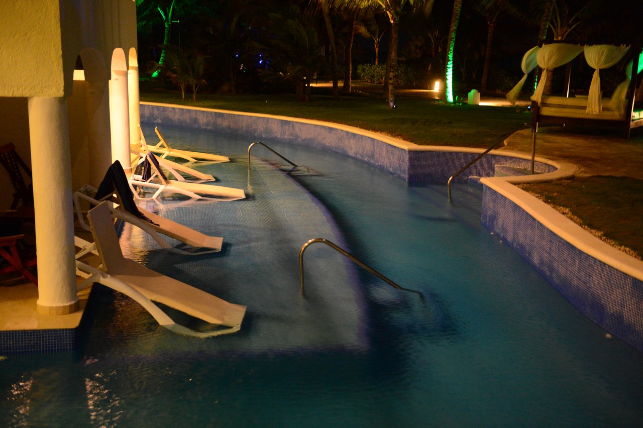 Several lounge chairs are partially submerged in a pool at night, with dim lighting and palm trees in the background.