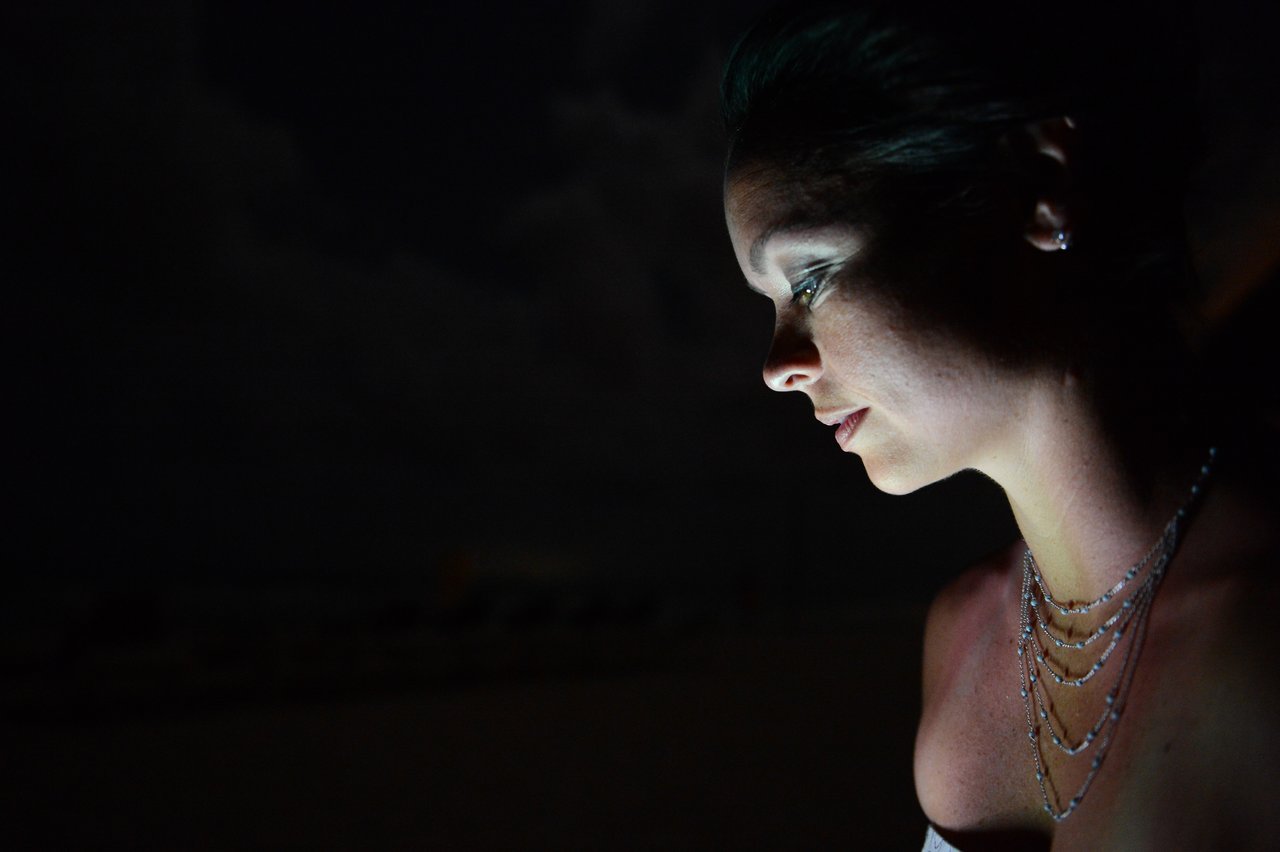 A woman in a necklace looks at a light source in the dark, her face softly illuminated.