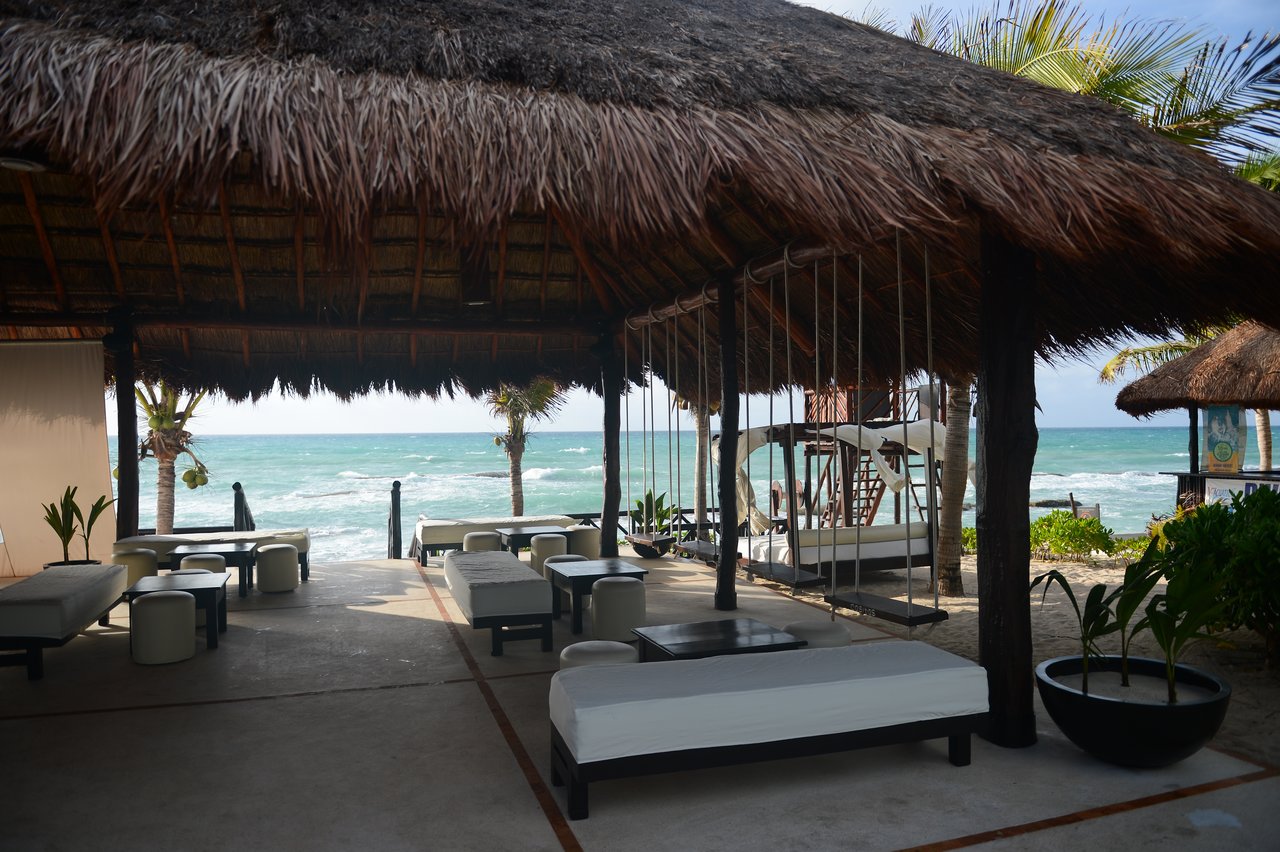 Open-air beachside lounge with wooden swings, cushioned seating, and ocean view under a thatched roof in Cancun.