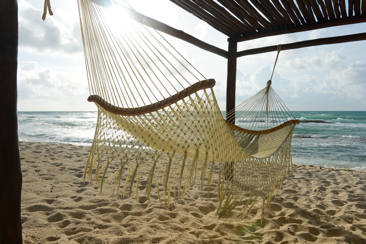 A woven hammock hangs under a wooden shelter on a sandy beach, with ocean waves in the background.