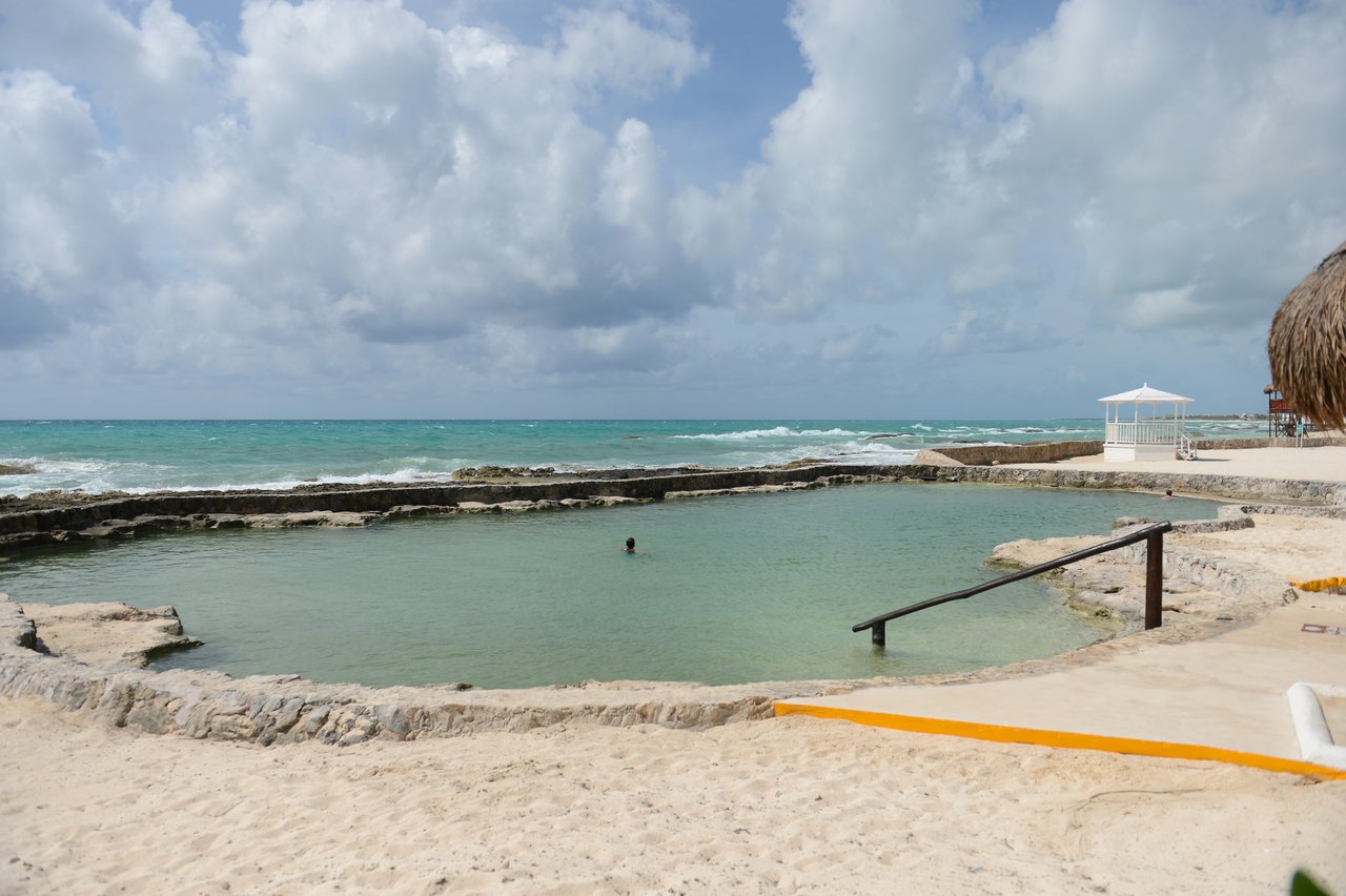 A person swims in a natural pool by the ocean, surrounded by stone edges and sandy beach.