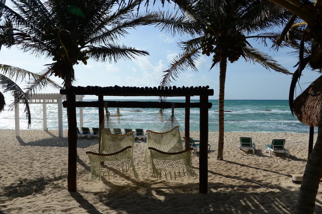 Two hammocks hang from a wooden frame on a sandy beach, facing the ocean with waves and lounge chairs nearby.