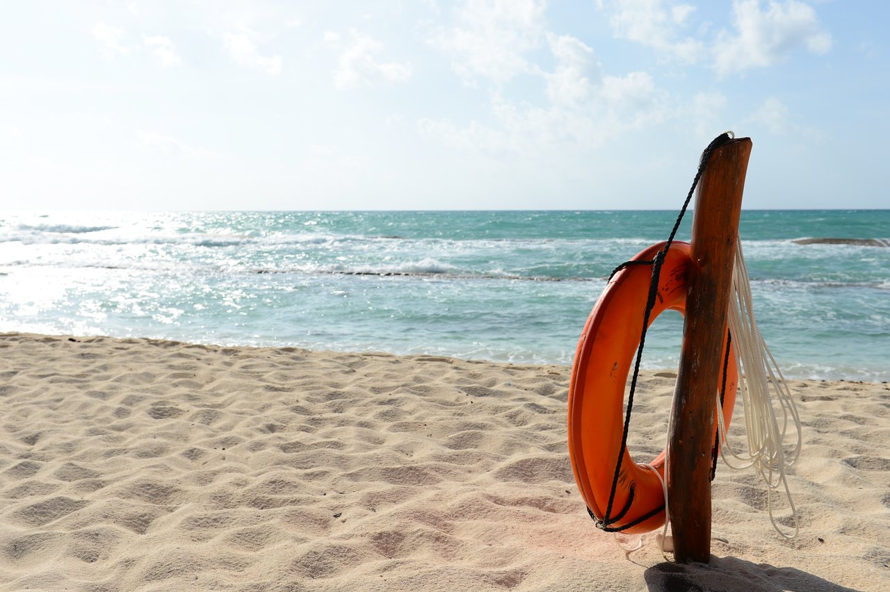 An orange lifebuoy hangs on a wooden post in the sand near the ocean waves.