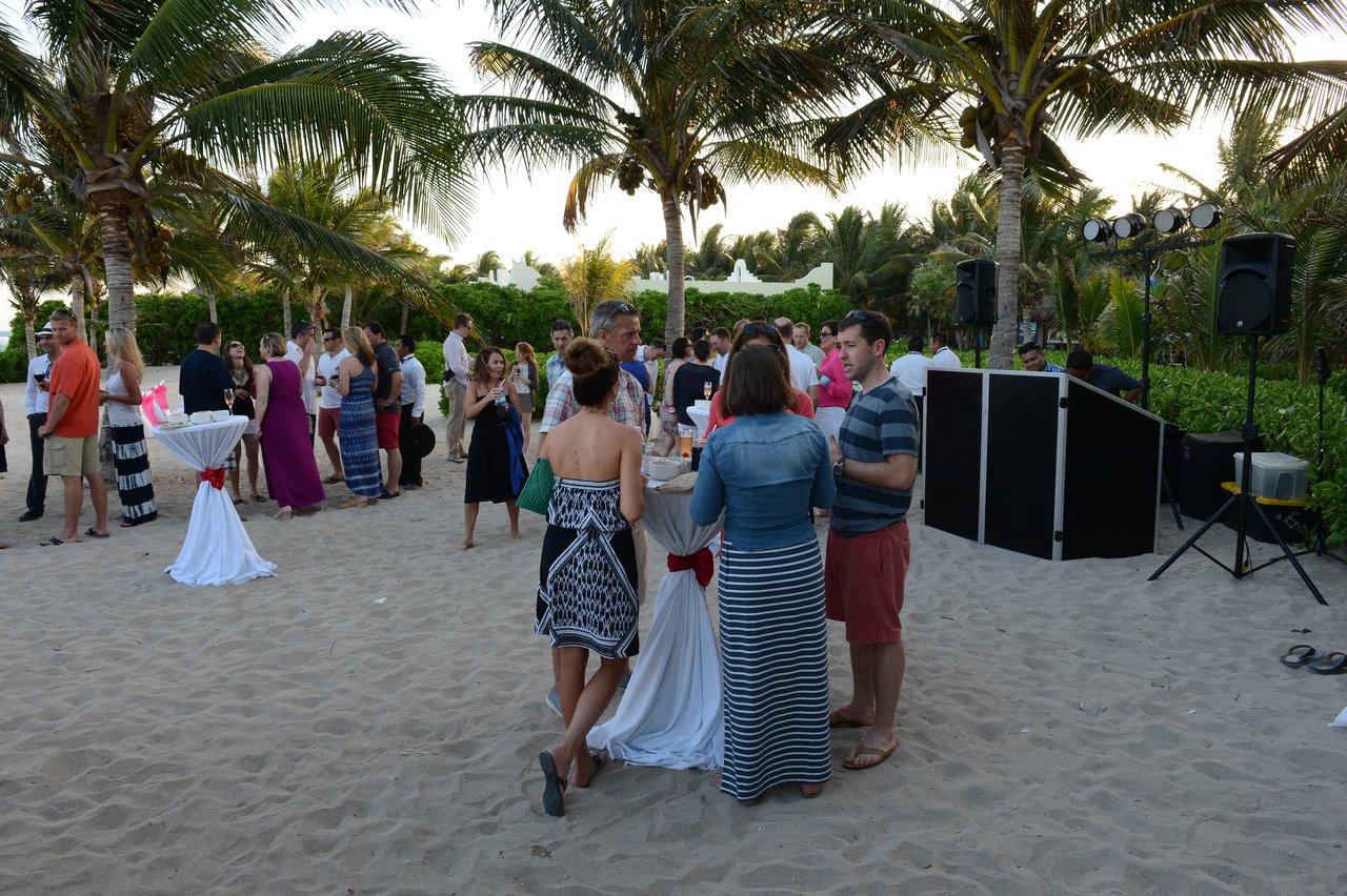People gathered on a sandy beach, socializing around cocktail tables with drinks, while a DJ setup is nearby.