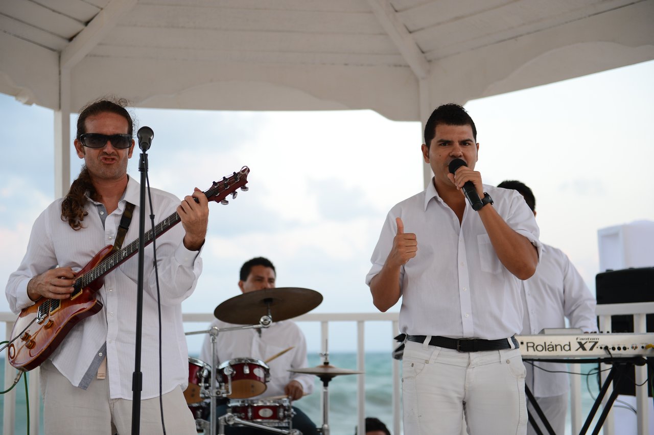 A band performs on an outdoor stage; a guitarist plays, and a singer holds a microphone while giving a thumbs-up.