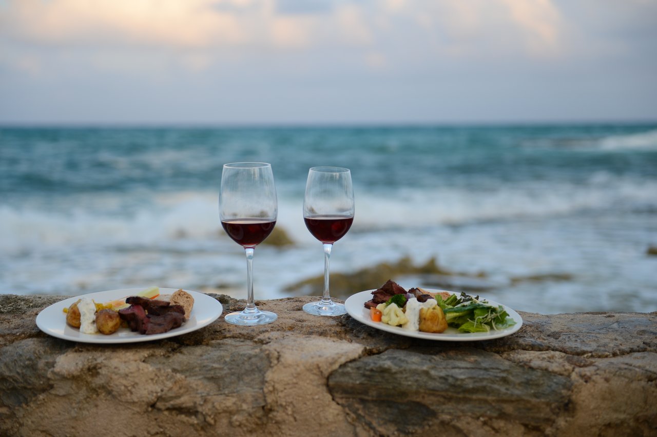 Two plates of food and two glasses of red wine on a stone ledge overlooking the ocean.