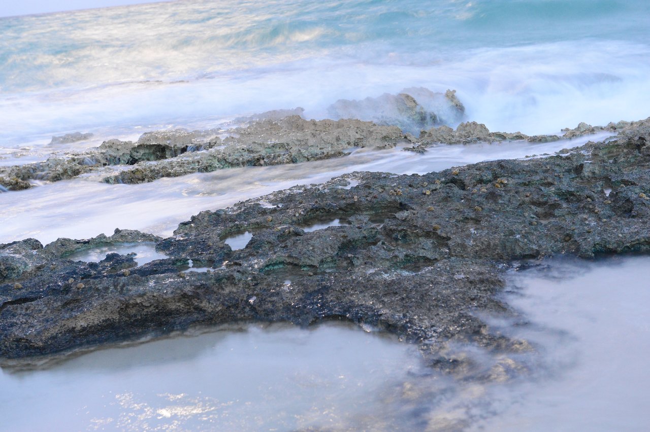 Waves washing over rugged, dark rocks on the shore, creating a misty effect in the water.