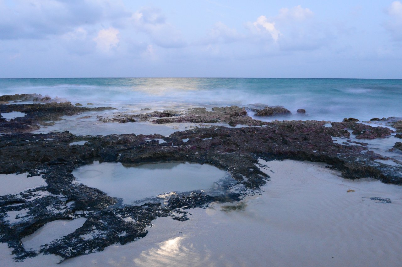 Rocky shoreline with shallow tide pools, waves gently washing over the rocks, and a cloudy sky in the background.