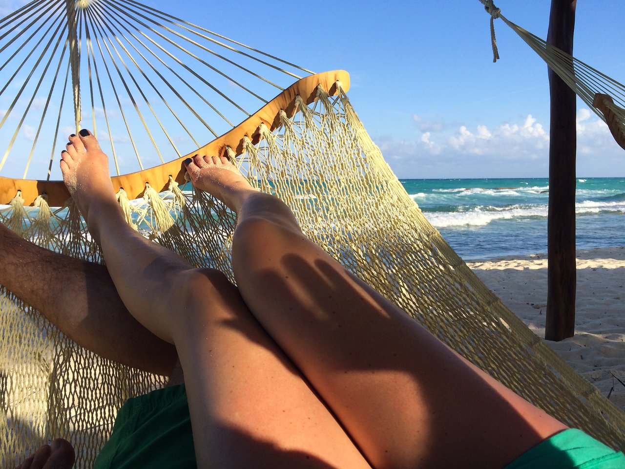 Two people relaxing in a hammock on a sandy beach, with ocean waves and a clear blue sky in view.