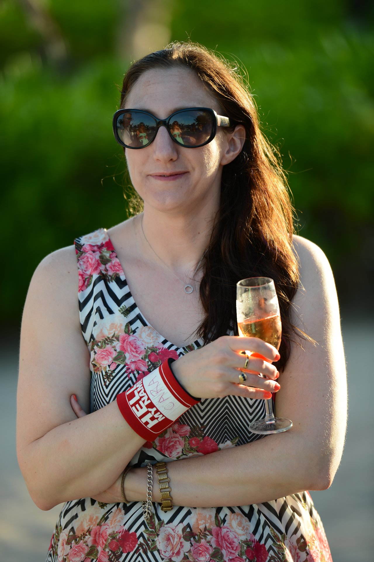 A woman wearing sunglasses and a floral dress holds a glass of champagne while standing outdoors.