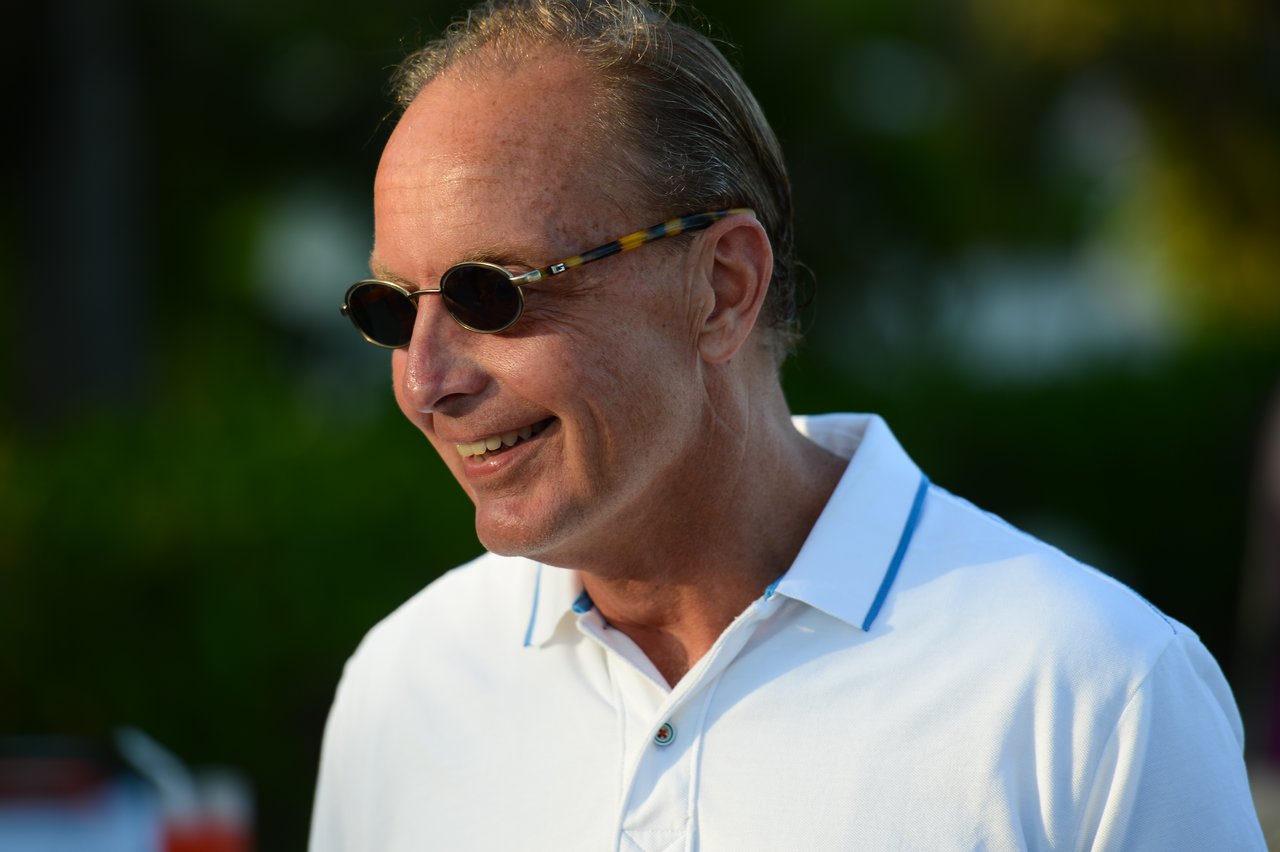 A man wearing sunglasses and a white polo shirt smiles while looking to the side outdoors.