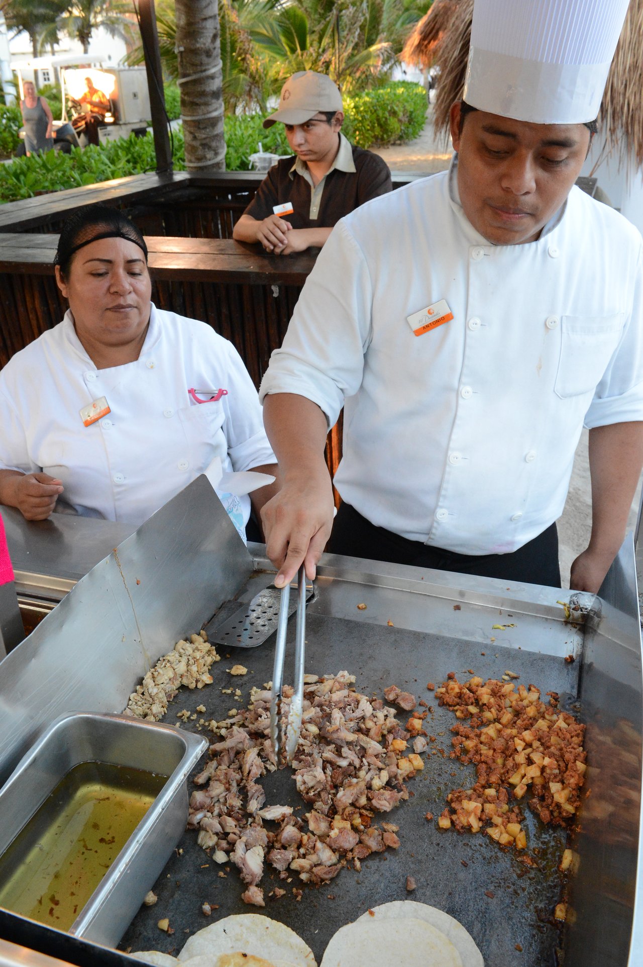 A chef in a white uniform prepares tacos on a grill, using tongs to handle chopped meat.