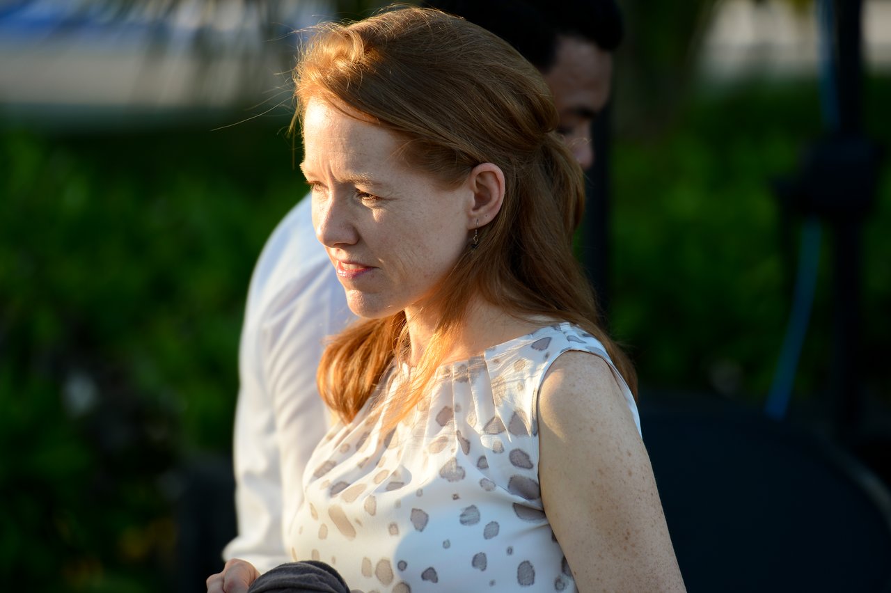 A woman with red hair stands in sunlight, looking to the side.
