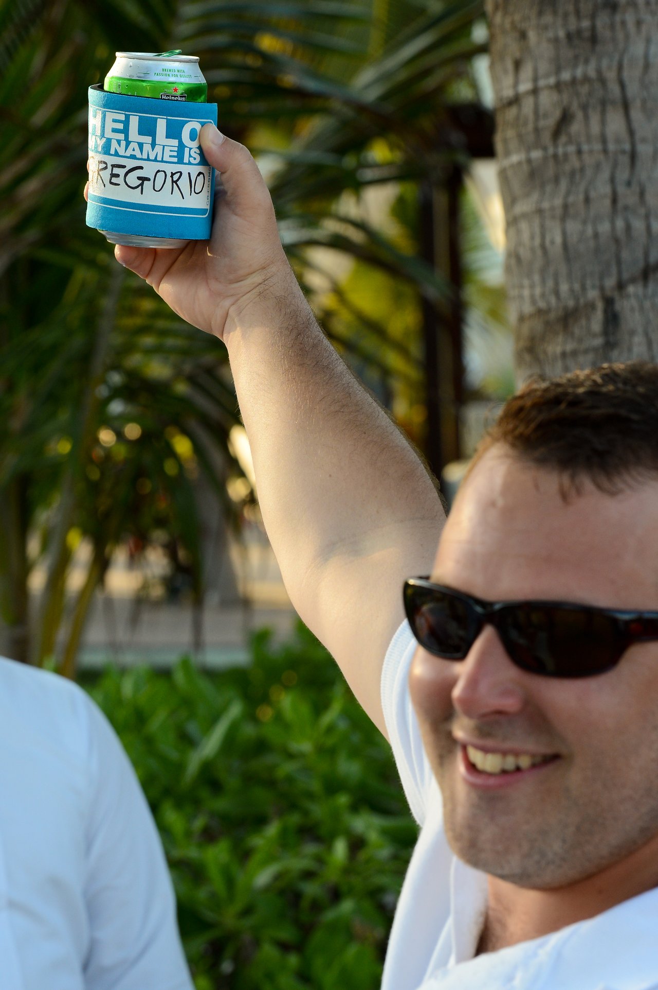 A man wearing sunglasses smiles while raising a drink in a blue holder labeled "Hello, my name is Gregorio.
