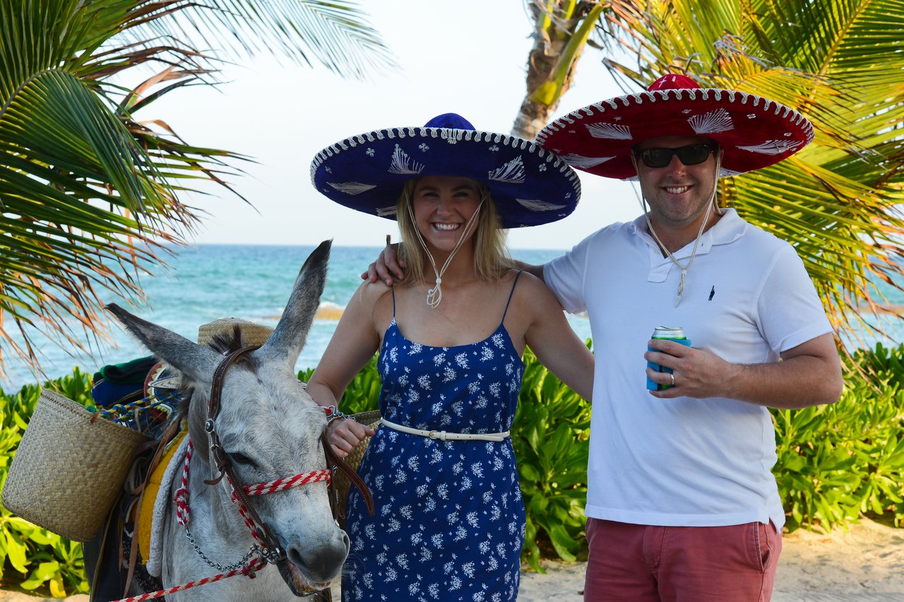 A smiling couple in sombreros poses with a donkey on a beach, with the ocean and palm trees behind them.