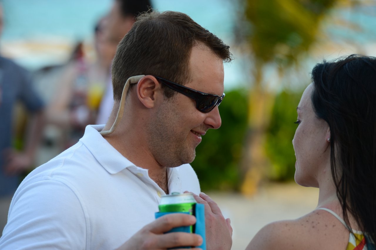 A man in sunglasses holds a drink and smiles while talking to a woman at the beach.