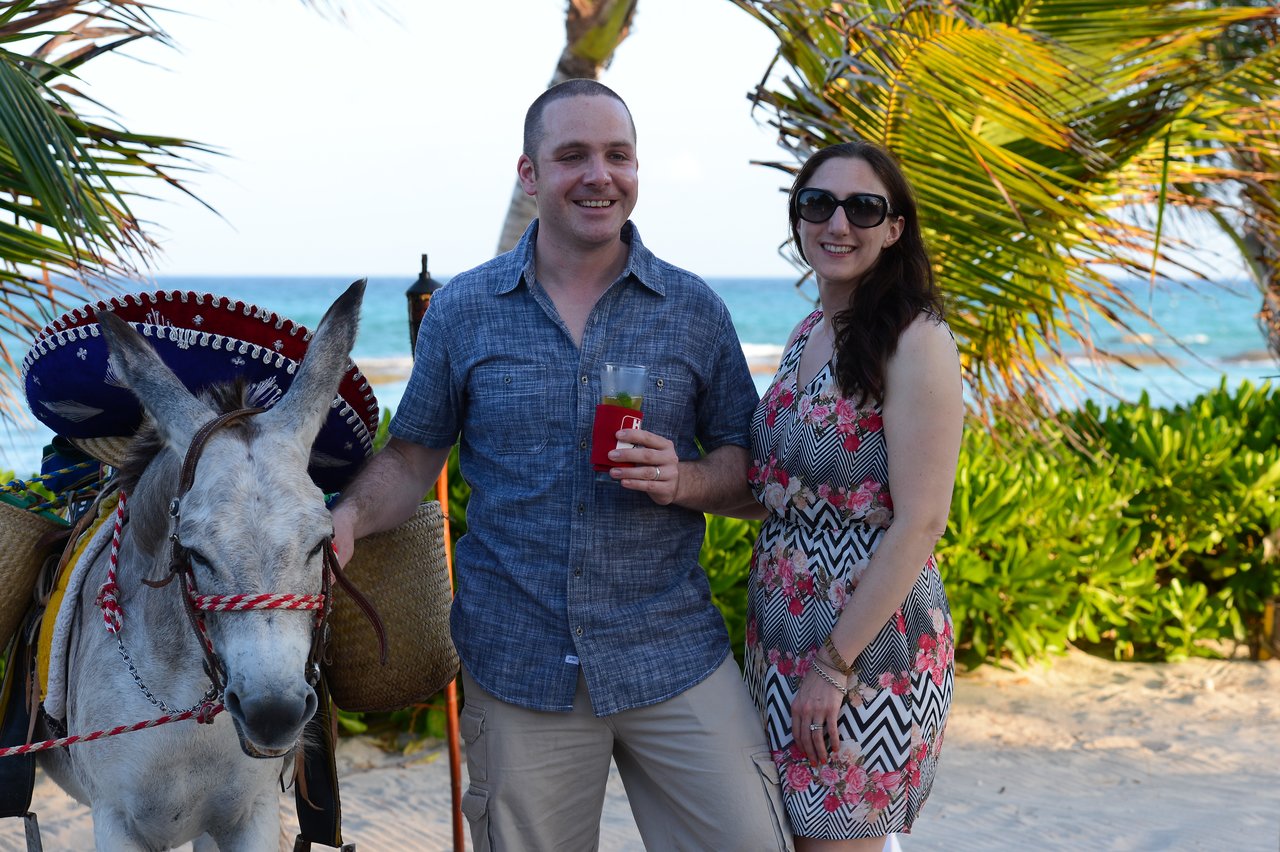 A man and woman smile at the camera on a beach, standing next to a donkey wearing a sombrero.