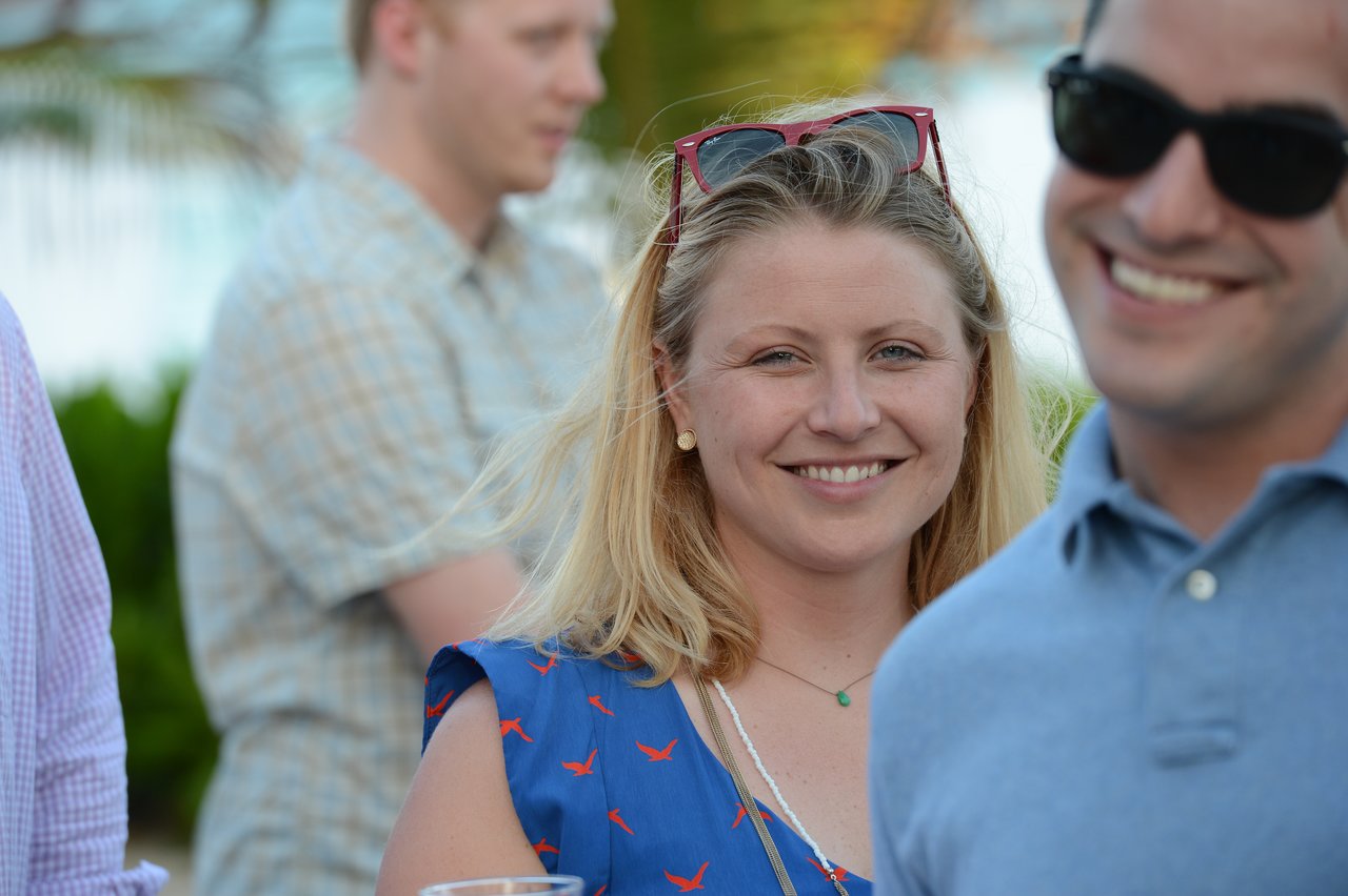 A smiling woman with sunglasses on her head looks at the camera, while others socialize in the background.