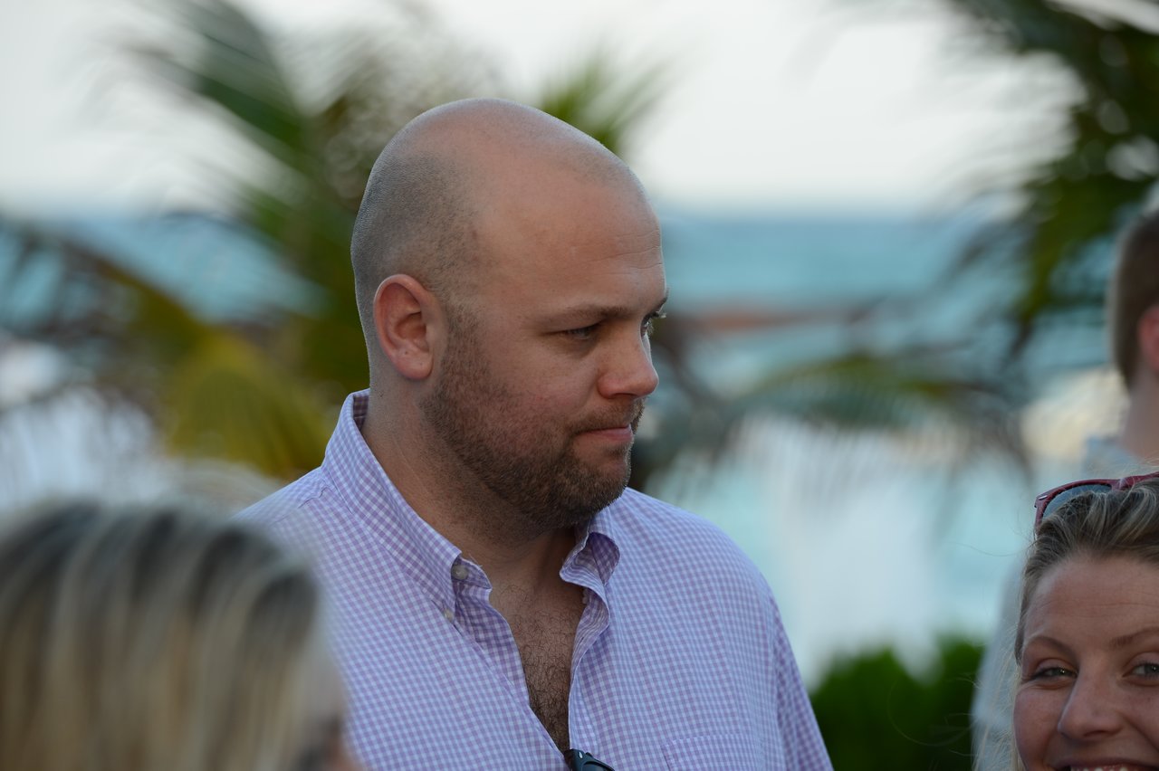 A man in a light purple shirt looks to the side while standing outdoors with others near the beach.