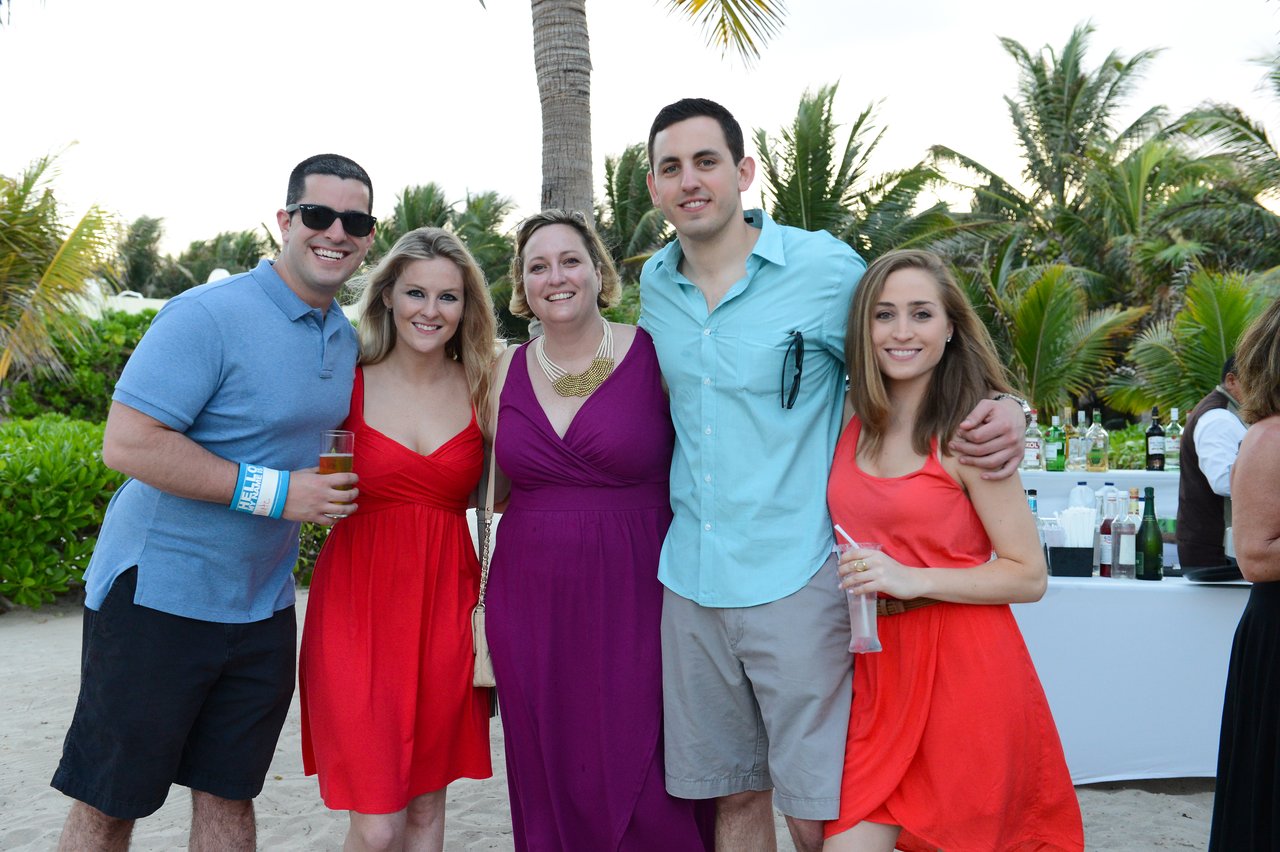 Five people posing together at an outdoor event on a beach, dressed in casual and semi-formal attire, holding drinks.
