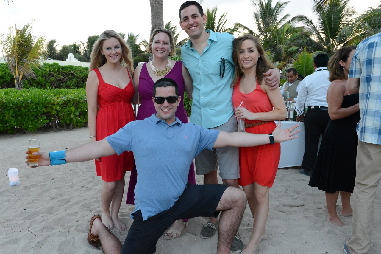 A group of five people poses on a sandy beach, with one man crouching in front with arms outstretched.