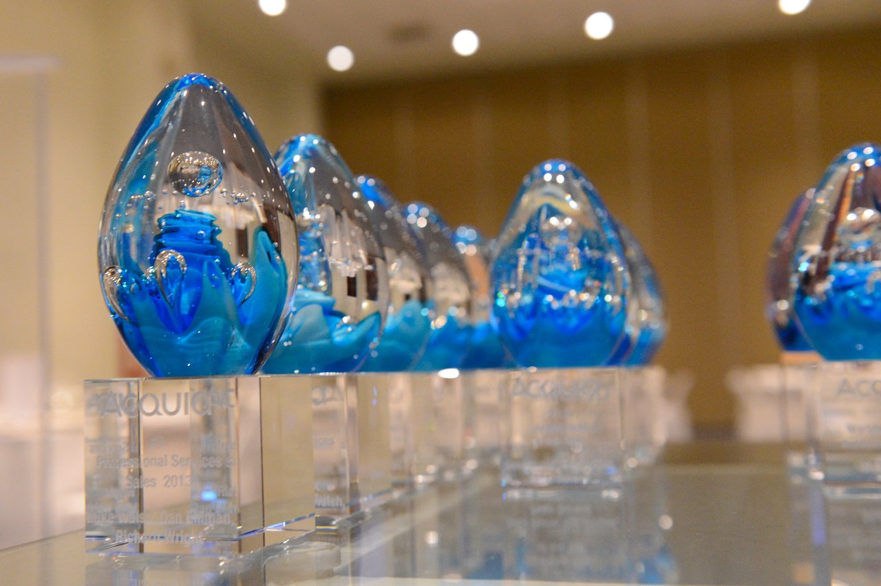 A row of blue glass awards with engraved bases displayed on a reflective table in a well-lit room.