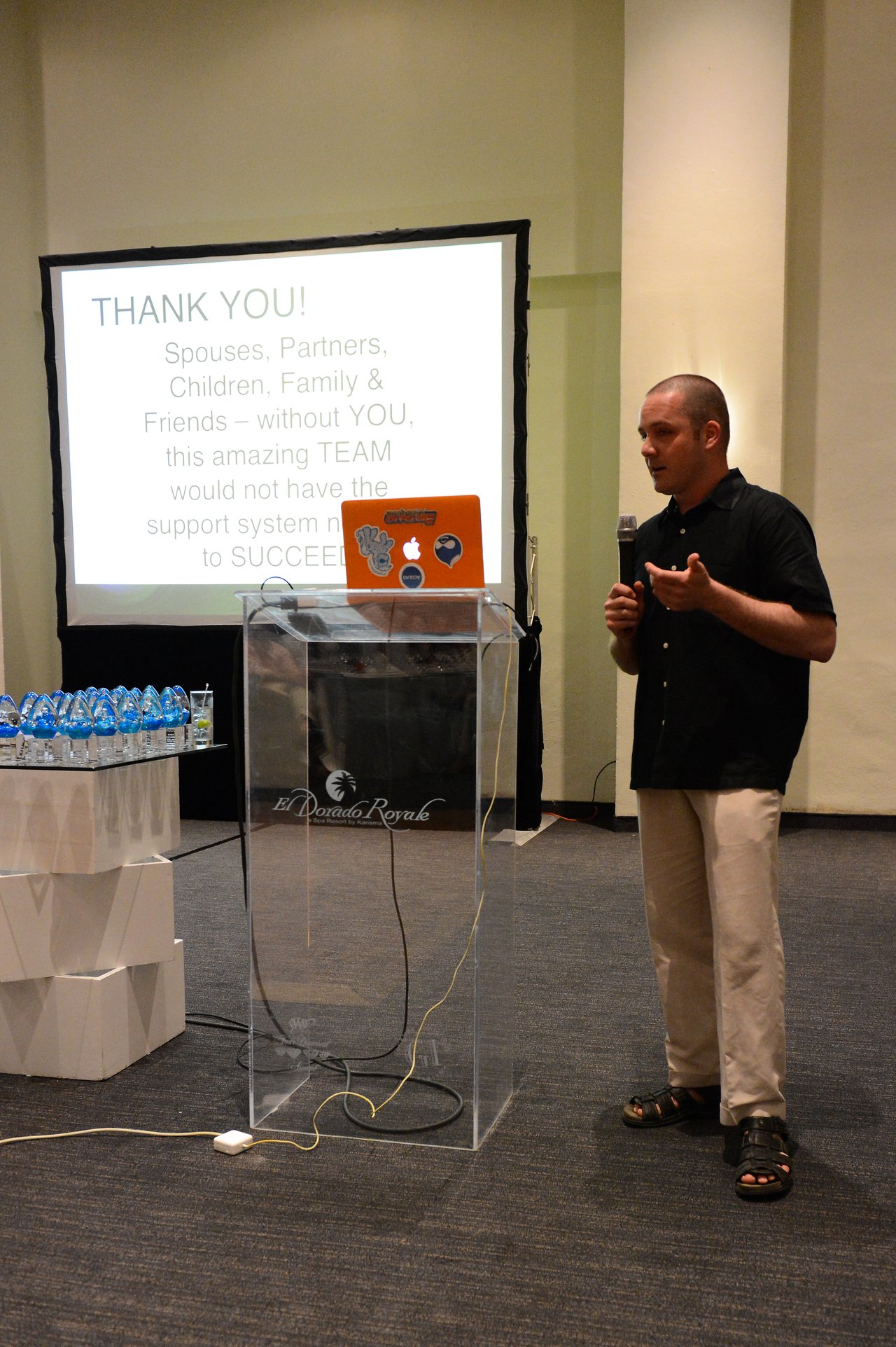 A man gives a presentation, holding a microphone near a podium with a laptop and a projection screen behind him.