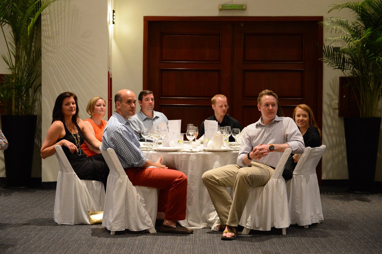 A group of people sits at a round table in a banquet hall, attentively watching something off-camera.