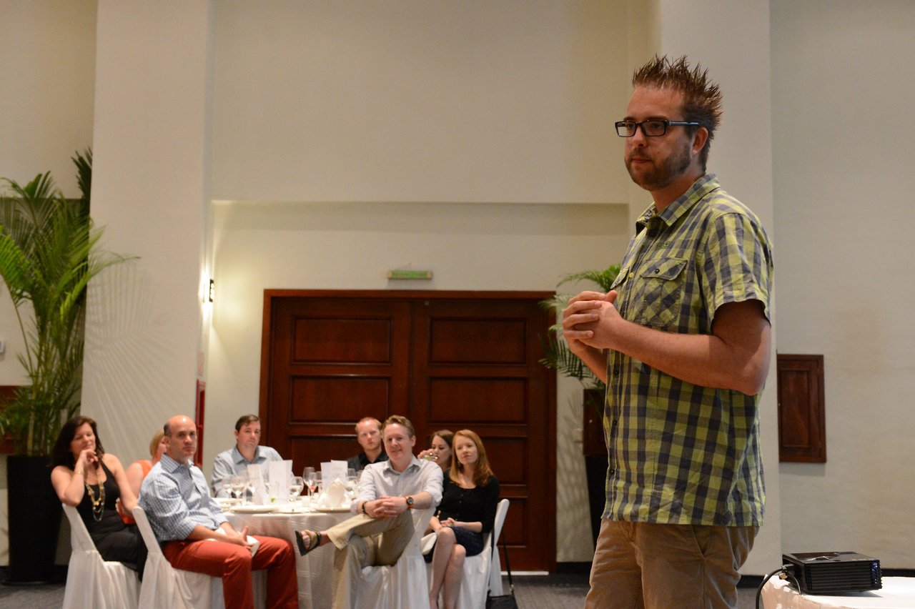 A man in a plaid shirt speaks to an audience seated at round tables in a conference room.