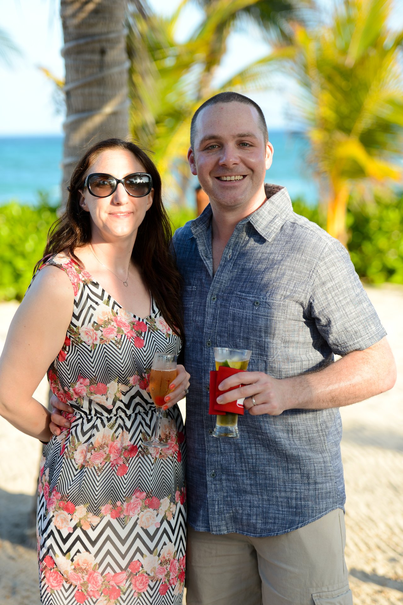 A smiling couple poses outdoors in Cancun, holding drinks, with palm trees and the ocean in the background.