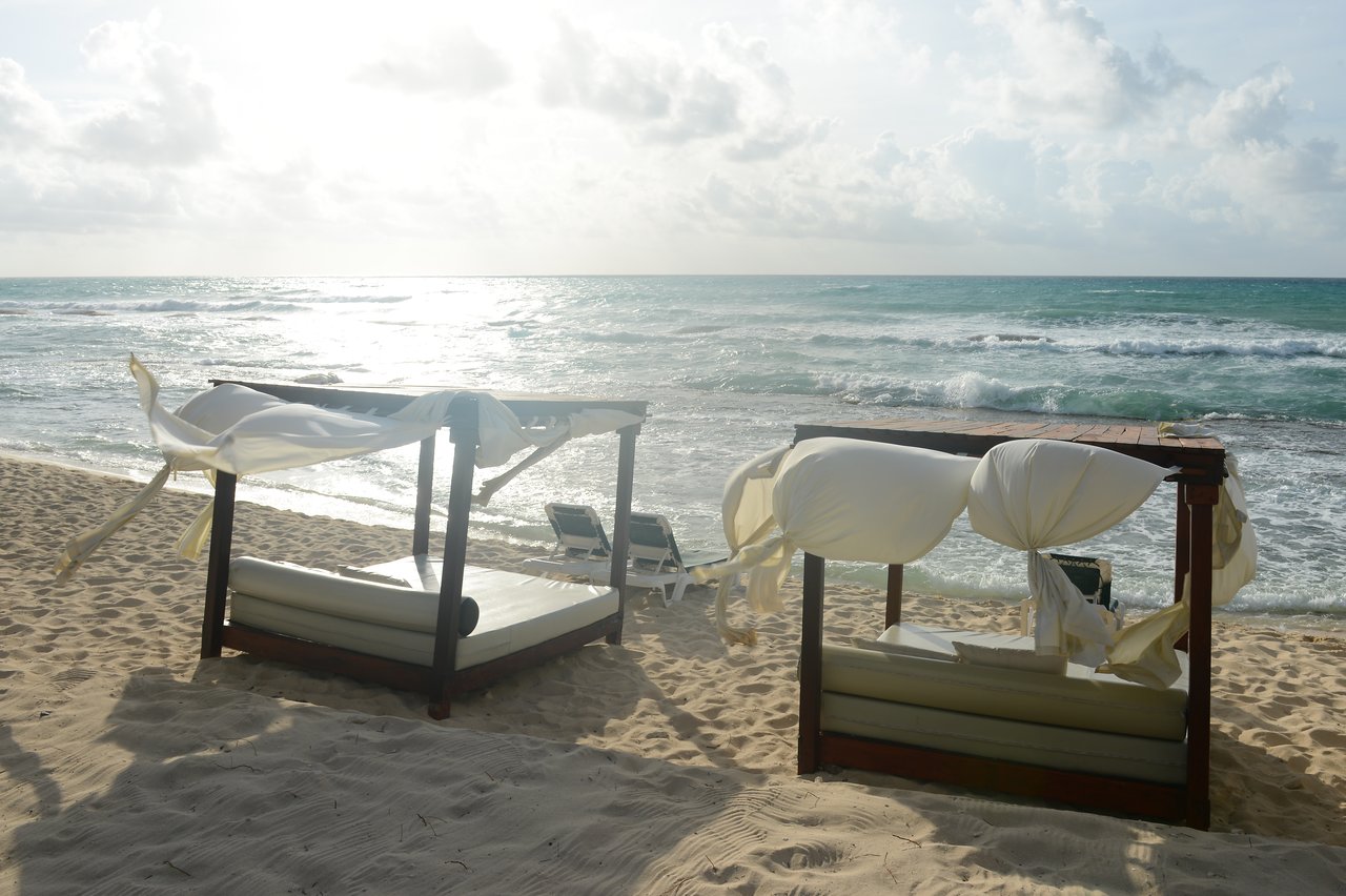 Two wooden beach beds with white cushions and flowing fabric face the ocean waves on a sandy shore.