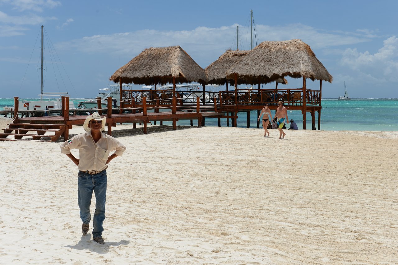 A man in a cowboy hat stands on a sandy beach, while two people walk nearby toward the water.