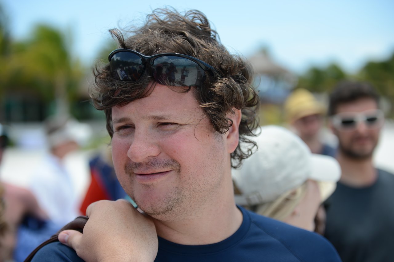 A man with curly hair and sunglasses on his head smiles while standing outdoors with a group of people.