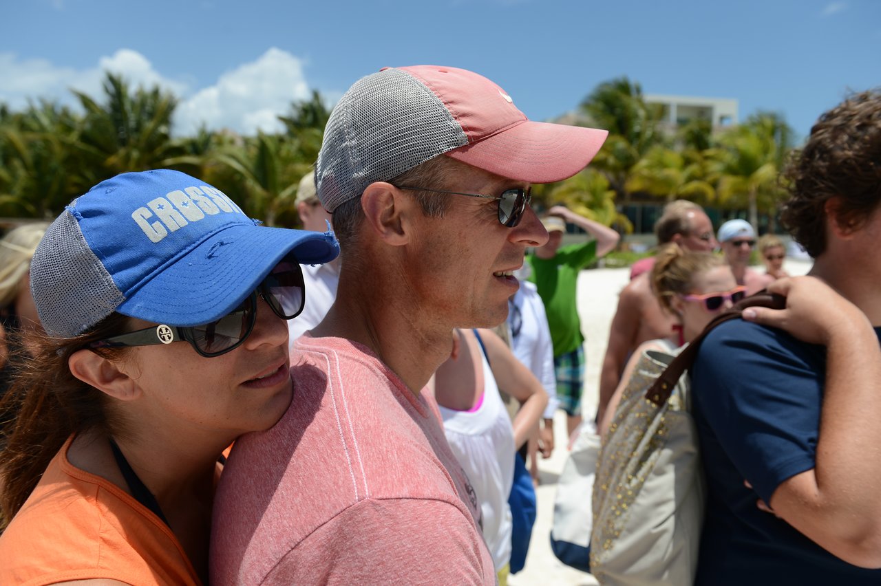 A woman in a blue cap leans on a man in a pink cap as they stand in a crowd on the beach.