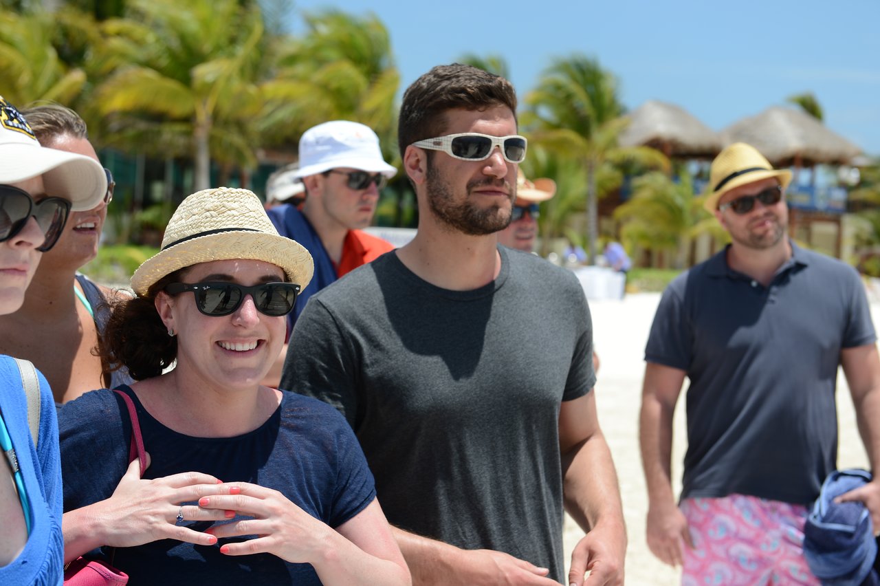 A group of people wearing sunglasses and hats stand on a sunny beach, looking in the same direction.